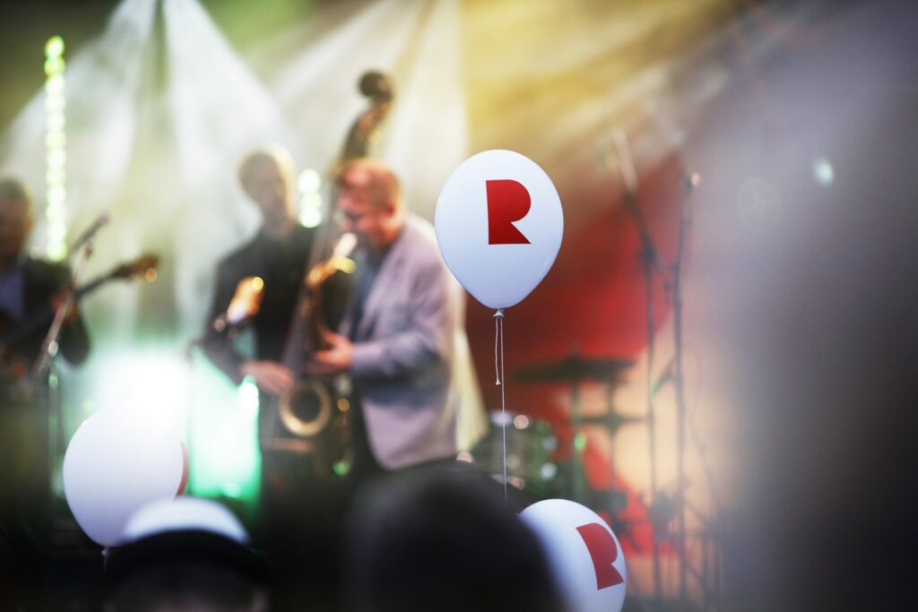 Musiikkikonsertti, jossa taustalla mies soittaa saksofonia värikkäiden valojen loisteessa. Etualalla valkoinen ilmapallo, jossa punainen R-elementti.
