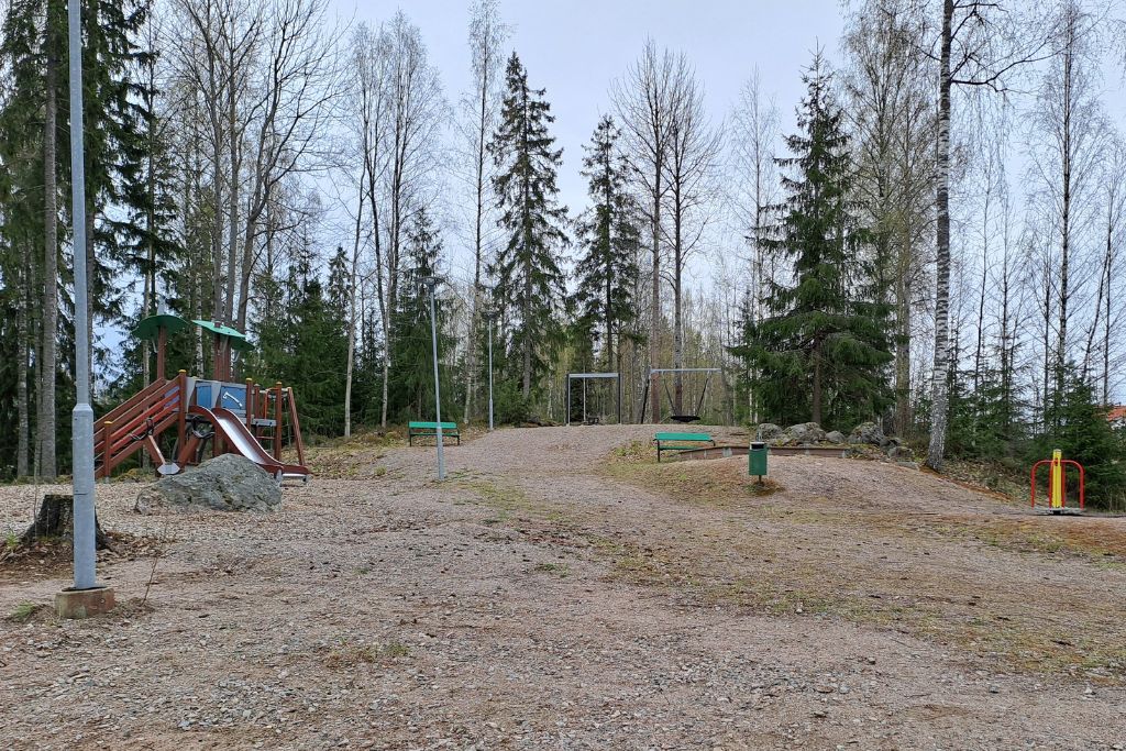 Hiekkamäki, jolla lasten leikkivälineitä, muun muassa liukumäki ja keinut. Taustalla metsää.