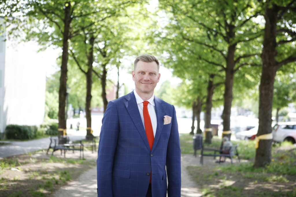Sinisessä puvussa oleva mies hymyilee ulkona puistoraitin/puukujan keskellä.