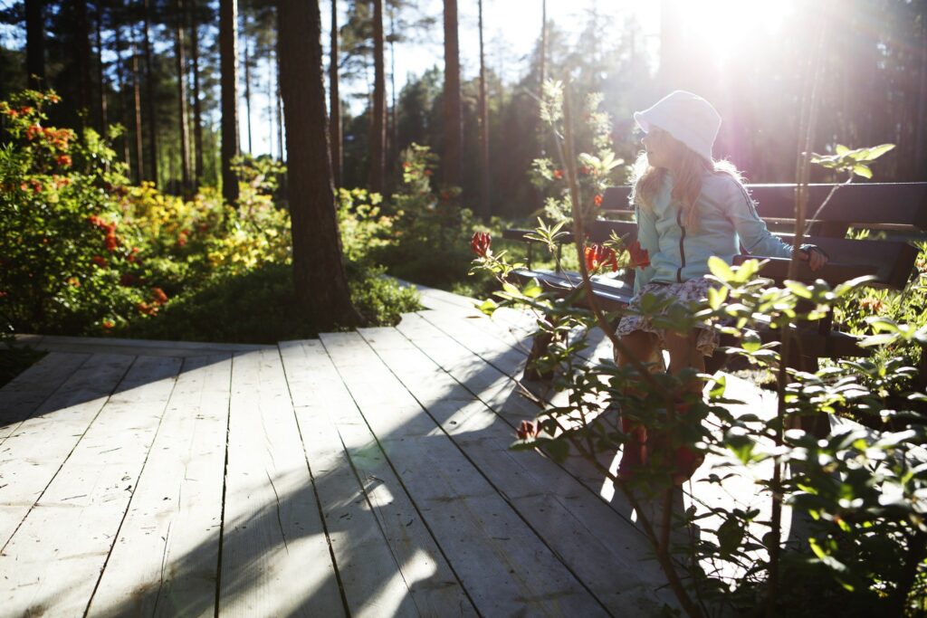 Vaaleahiuksinen tyttö istuu puisella puiston penkillä. Taustalla puita ja vehreää kasvillisuutta ja kukkia.