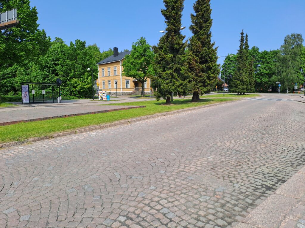 Mukulakivikatua kaksi tietä vierekkäin, välissä nurmikkokaistale. Kauemman kadun varrella bussipysäkki, taustalla puita ja keltainen puurakennus.