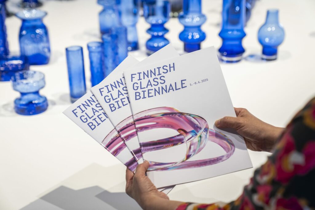 Henkilöllä käsissään viuhka esitteitä, joiden kannessa lukee Finnish Glass Biennale. Taustalla sinisiä lasimaljakoita pöydällä.