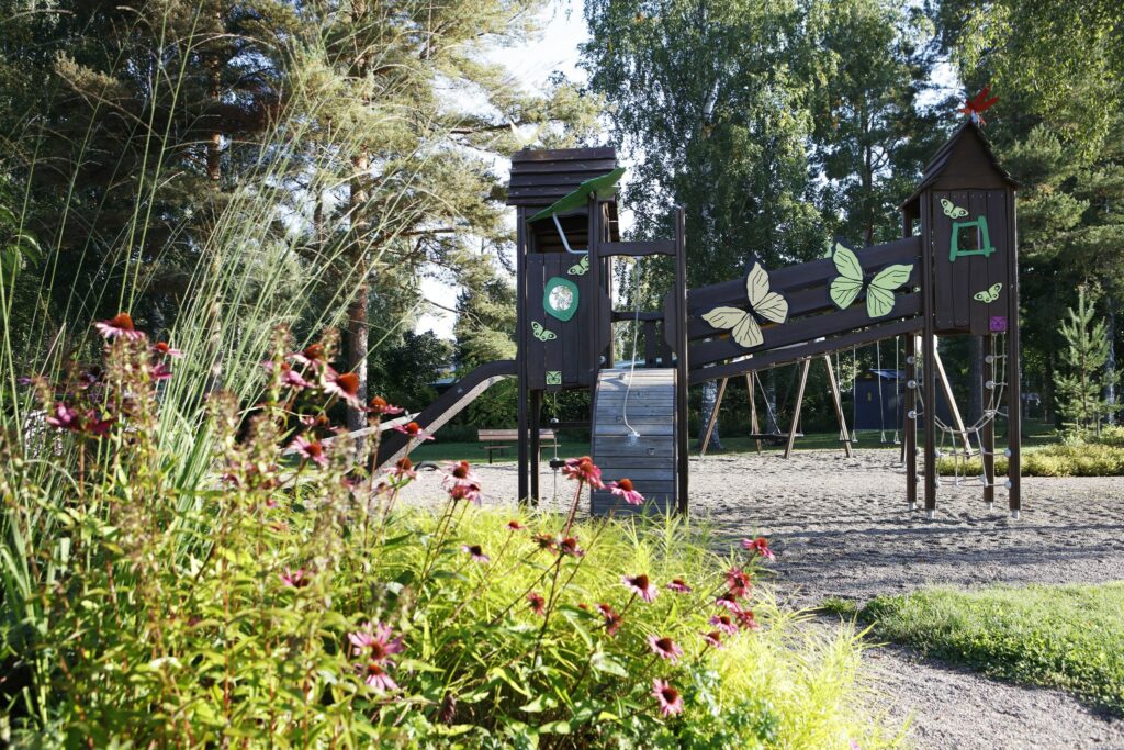 Etualalla kasvillisuutta ja taaempana leikkipuiston kiipeilyteline isoilla perhoskoristeilla