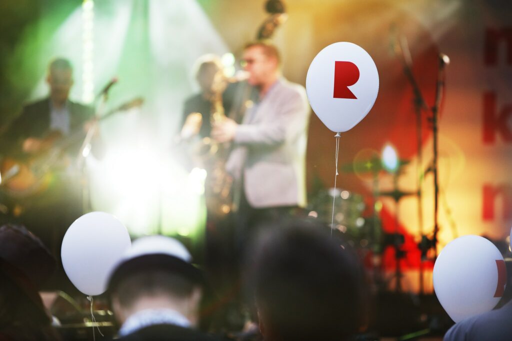Yleisön yllä leijuu ilmapallo, jossa on Riihimäen kaupungin R-tunnus. Taustalla yhtye soittaa musiikkia.