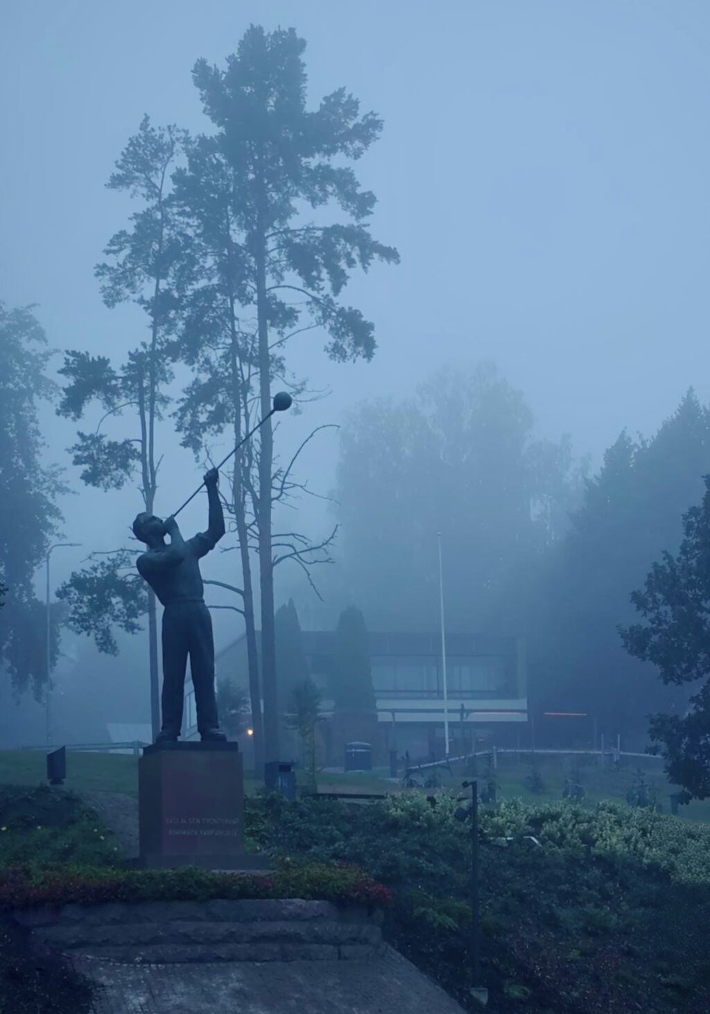 Utuisen sininen kuva, jossa etualalla on lasinpuhaltajapatsas ja taustalla puita sekä rakennus