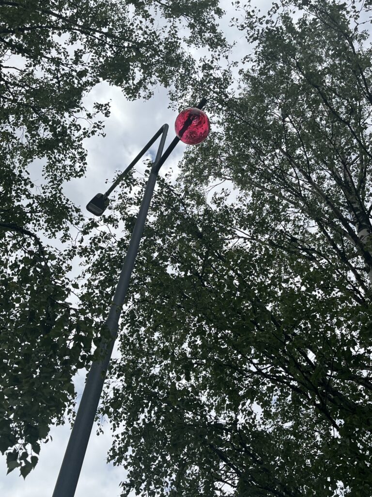 Alhaaltapäin otettu kuva katulampusta, jonka päässä on punainen lasipallo. Ympärillä puita.