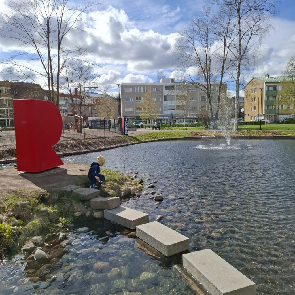 Puistoalue, jossa on punainen R-teos, lampi ja pieni lapsi tutkimassa lampea.