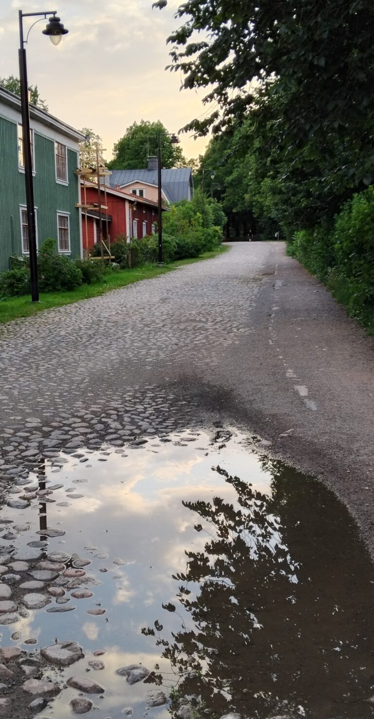 Mukulakivi, jossa iso vesilätäkkö. Tien vierustalla puita ja vanhoja puutaloja.