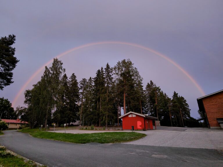 Puita ja rakennuksia iltamaisemassa. Keskellä taivaalla suuri sateenkaari.