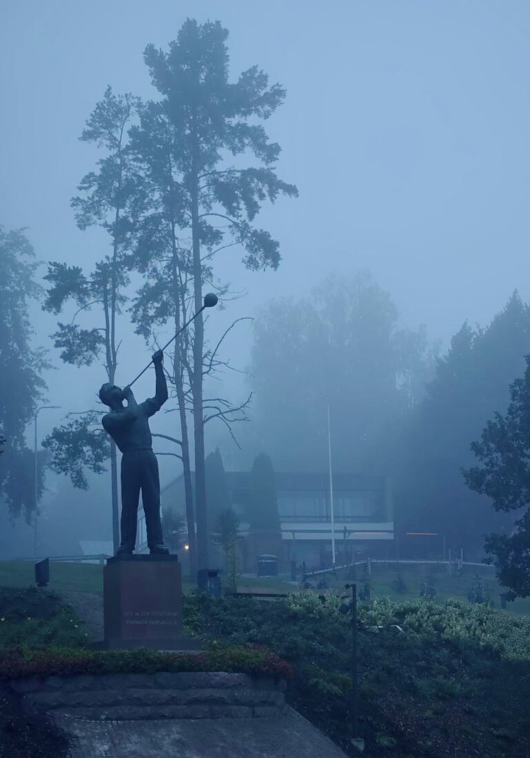 Utuisen sininen kuva, jossa etualalla on lasinpuhaltajapatsas ja taustalla puita sekä rakennus