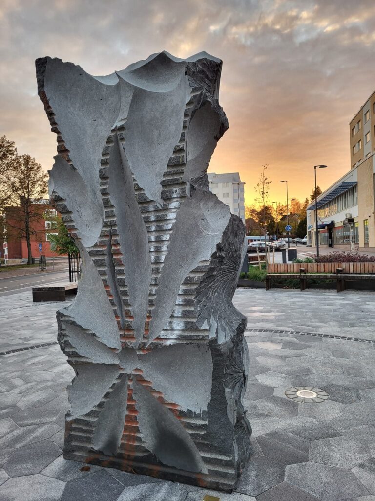 Harmaa abstrakti ja jykevä veistos keskellä kaupunkia auringonkajossa.