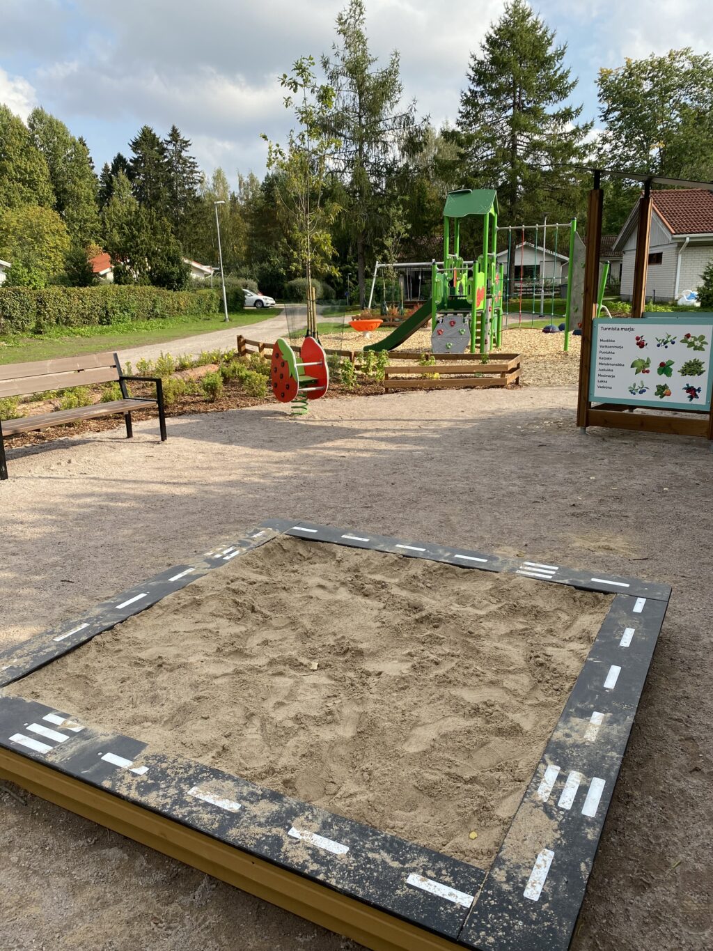 Etualalla hiekkalaatikko, jonka reunoihin on tehty ajorata. Taaempana näkyy mansikka-kiikku ja vihreä kiipeilyteline.