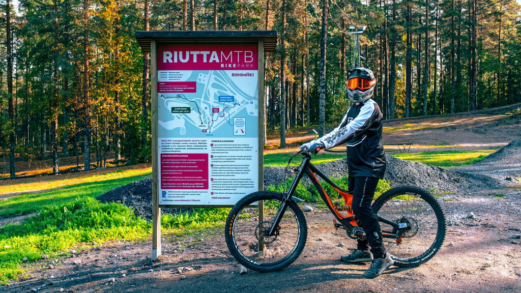Ihminen, jolla on koko kasvot peittävä kypärä päässään, pyöränsä kanssa Riutta MTB-opastekyltin luona ulkona metsämaastossa.