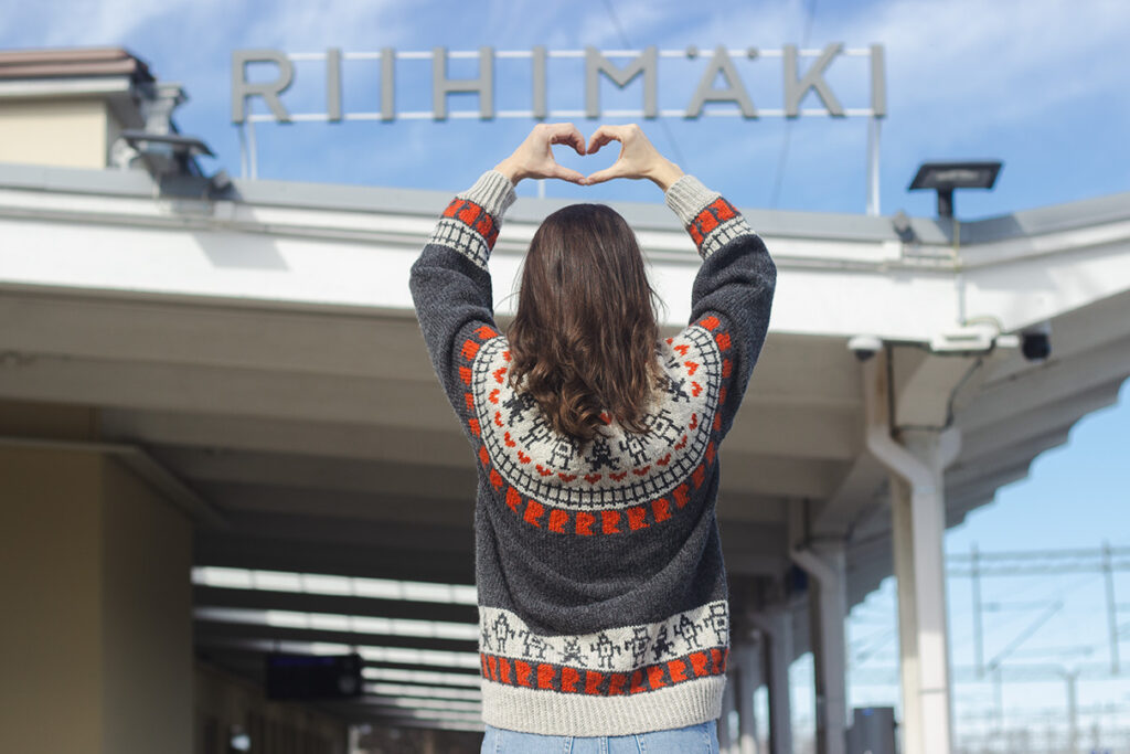 Riihimäki-villapaita takaapäin kuvattuna ruskeatukkaisen naisen päällä. Nainen seisoo villapaita päällä Riihimäen juna-aseman edessä ja hänellä on kädet pään yläpuolella muodostaen sormillaan sydämen.