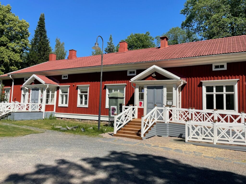 Punainen pitkä puurakennus, jossa kaksi sisäänkäyntiä. Hiekkapiha edessä, pilvetön taivas taustalla.