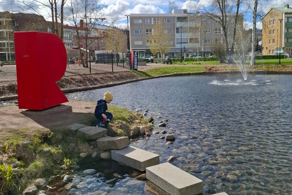 Muutaman vuoden ikäinen lapsi ulkoilemassa puistossa, jossa on suuri lampi. Lammen poikki johtaa harkoista tehty "kävelypolku". Taustalla puistossa punainen R-kirjaimen muotoinen veistos.