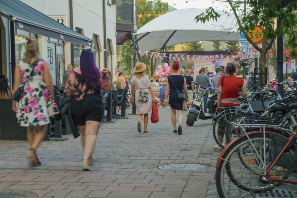 Ihmisiä kävelemässä kaupunkikeskustassa kadulla. Ihmisillä kesäinen vaatetus. Suuri kankainen katos ja värikkäitä viirejä kadun päädyssä, oikealla pyöräteline, jossa pyöriä.
