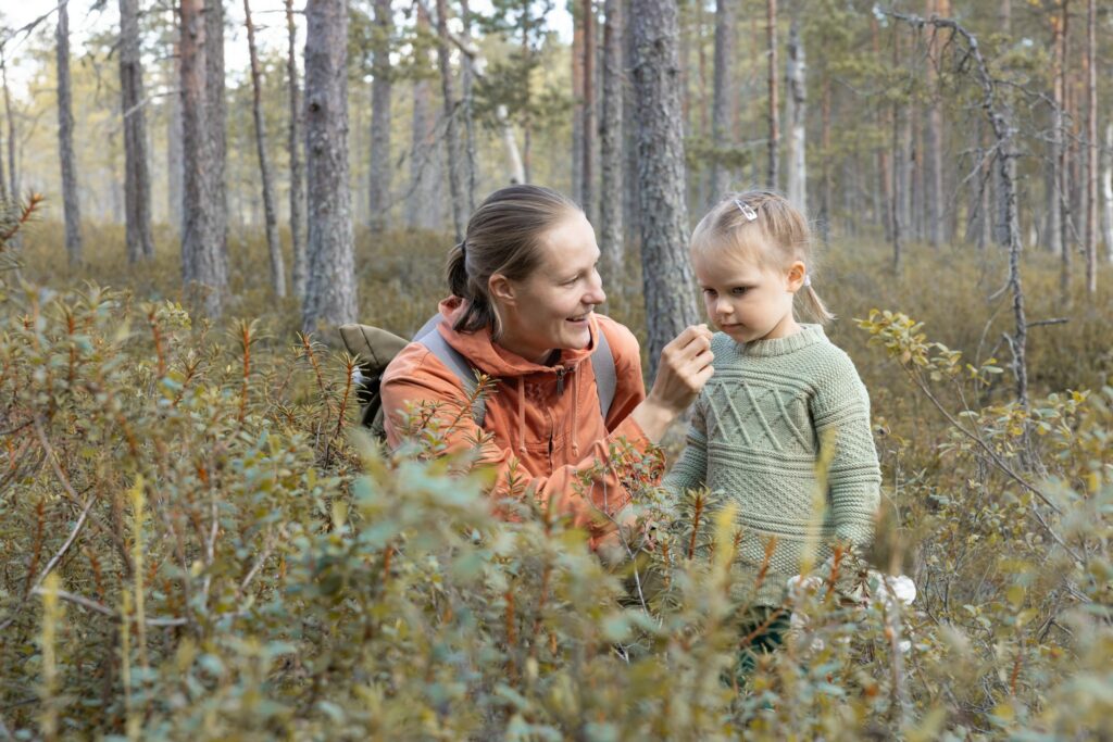 Nainen ja lapsi metsässä varvikossa, taustalla mäntyjen runkoja Nainen on kyykistynyt lapsen tasolle, näyttää lapselle jotain sormissaan olevaa. Syksyinen tunnelma.