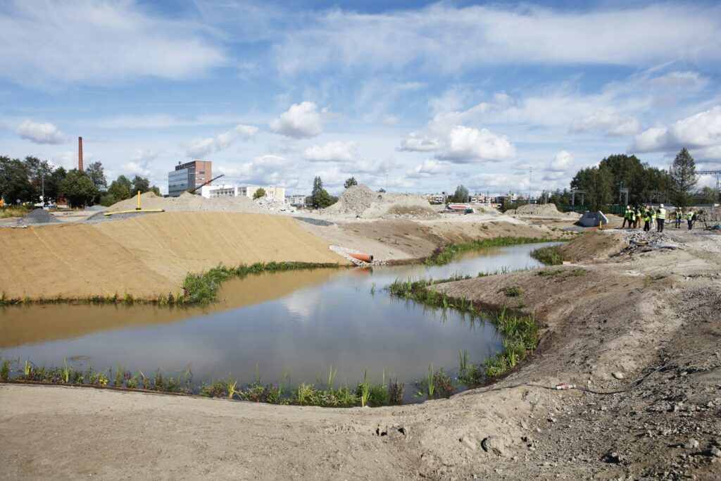 Mutkitteleva joen uoma, rakennetut vaaleat hiekkapenkereet. Taustalla rautatie. Vaaleansininen taivas, jolla valkoisia pilvenhattaroita.