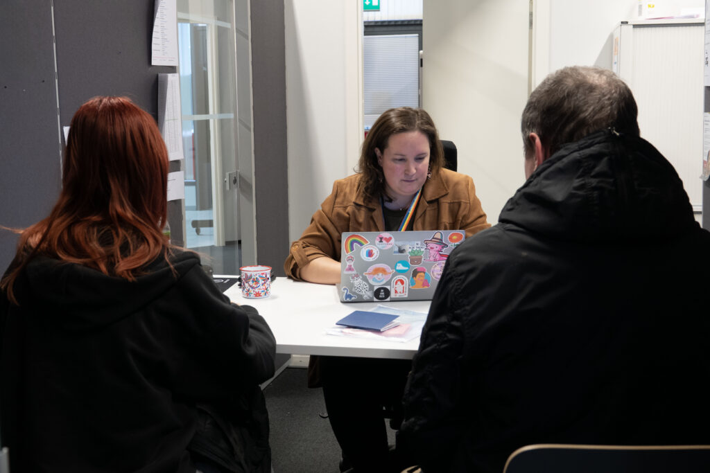 Yksi henkilö tietokoneen kanssa, kaksi henkilöä katsovat häntä.