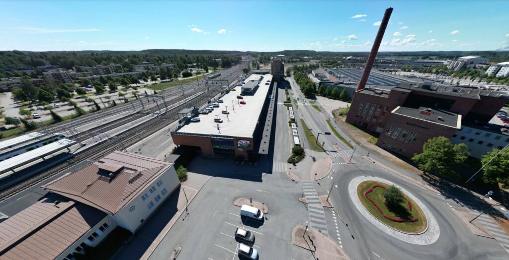 Viistoilmakuvanäkymä Rautatietorilta. Rautatie, pitkä matala matkakeskuksen rakennus sen kyljessä, liikenneympyrä ja autotie ja vanha voimalarakennus piippuineen. Kesäinen keskipäivä - aurinko paistaa lähes pilvettömältä taivaalta.