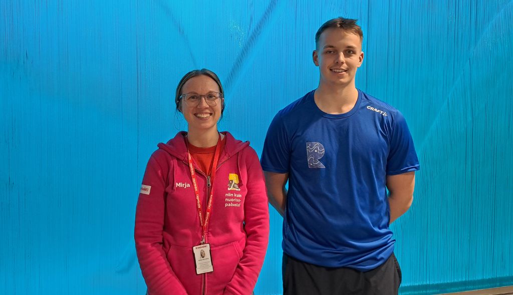 Nainen ja mies seisovat vierekkäin ja hymyilevät sinisen taustan edessä. Naisella punainen huppari ja miehellä sininen urhailuteepaita päällä.