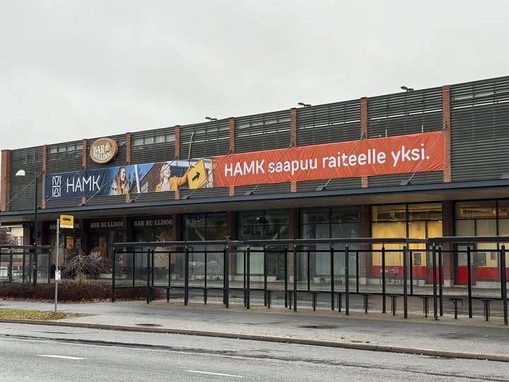 Autotie, jonka takana leveä rakennus, jonka kyljessä on iso mainoslakana, jossa lukee "HAMK saapuu raiteelle yksi". Harmaa taivas taustalla.
