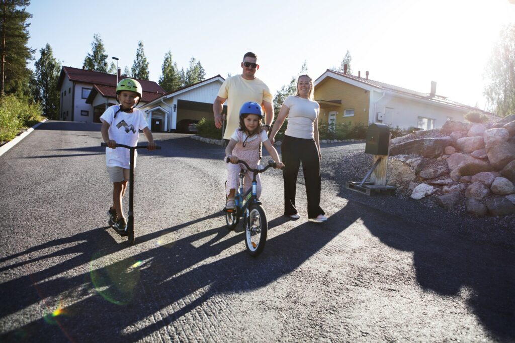 Äiti, isä ja kaksi lasta ulkoilemassa kesäisessä säässä. Toinen pienistä lapsista pyöräilee, toinen potkulautailee. Taustalla omakotitaloaluetta.