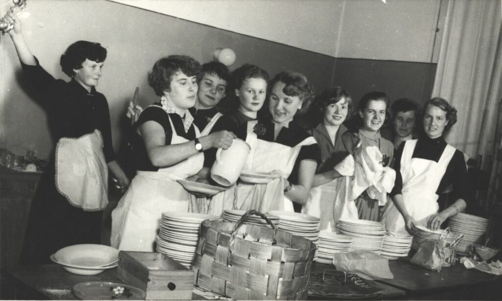 Mustavalkoinen valokuva, jossa yhdeksän hymyilevää naista essut päällä keittiön pöydän takana. Yksi on kaatavinaan kannusta toisen pitelemään astiaan. Pöydällä lautaspinoja. Kaikilla laitetut, kiharretut hiukset.