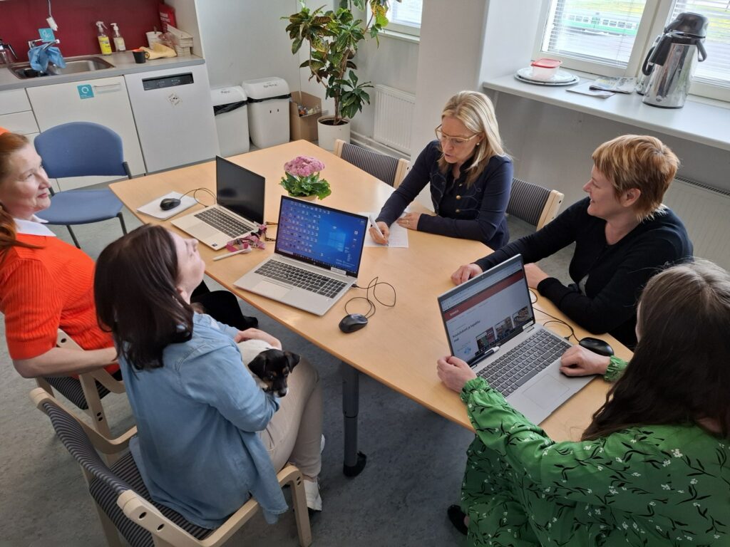 Viisi naista istumassa ja keskustelemassa toimistopöydän ääressä läppärit auki pöydällä.