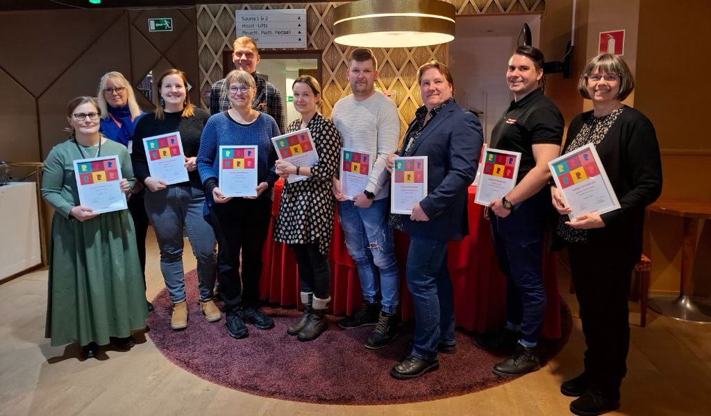 Kahdeksan henkilöä kunniakirjat käsissään rivissä sisällä salissa. Heidän taustallaan kaksi henkilöä ilman kunniakirjoja.