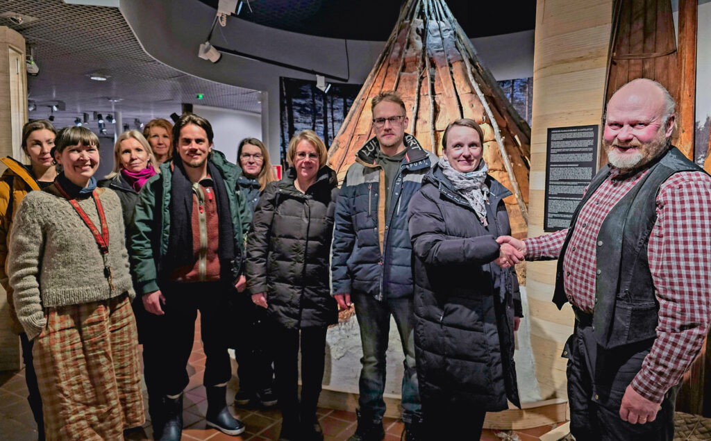 Noin kymmenen ihmistä koolla sisällä museossa. Kaksi heistä etualalla puristavat kättä. Muutkin hymyilevät kameralle. Taustalla sisältäpäin valaistu tiipiin näköinen teltta.