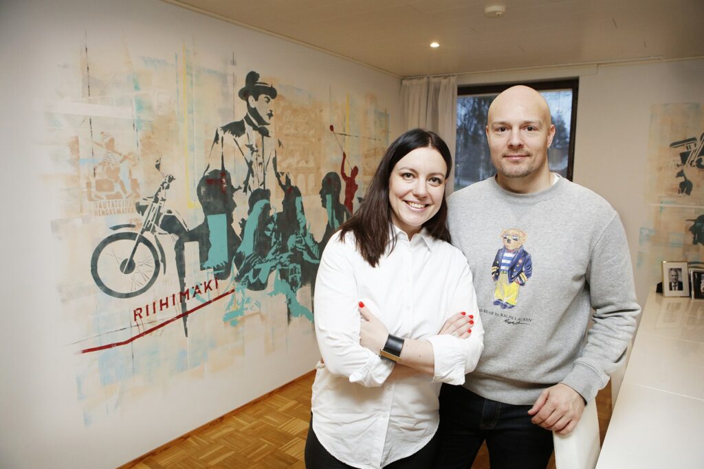 Nainen ja mies seisovat yhdessä hymyillen avarassa valoisassa huoneessa seinälle maalatun värikkään muraalin edessä.