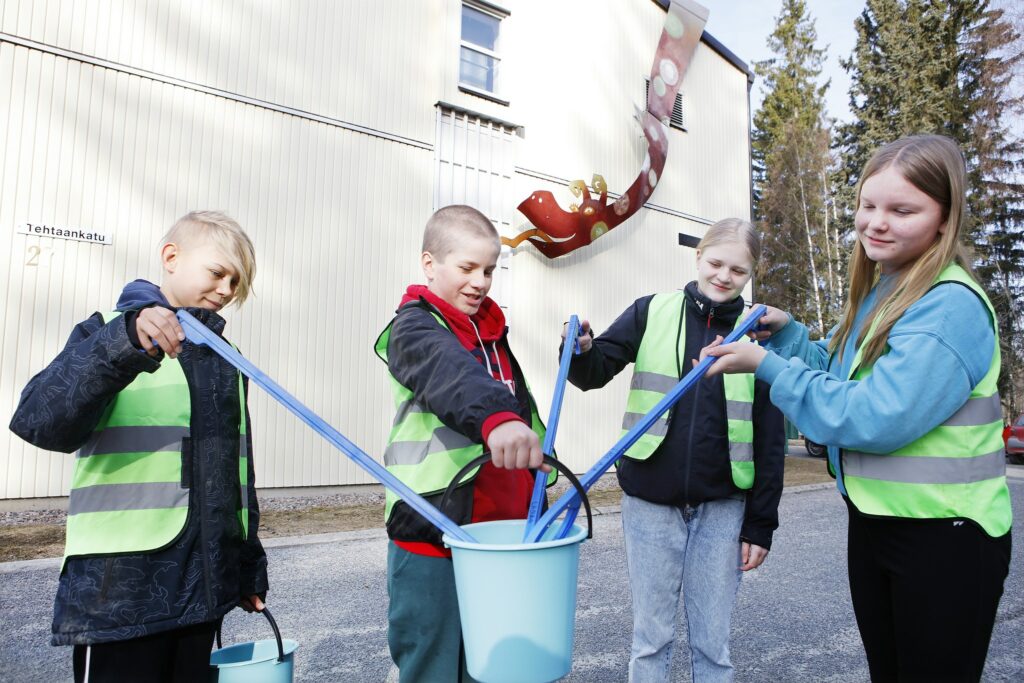 Neljä lasta koulun sivustalla ulkona heijastinliivit päällään laittamassa ämpäriin roskapihdeillä roskaa, kaikki neljä yhtäaikaa.