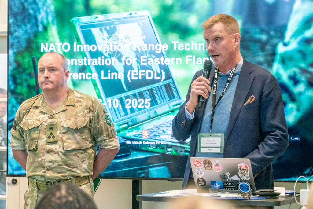 Mies puhumassa kokoussalissa mikrofoniin esitys ruudulla taustallaan. Vieressä seisoo mies, jolla on maastopuku päällään.