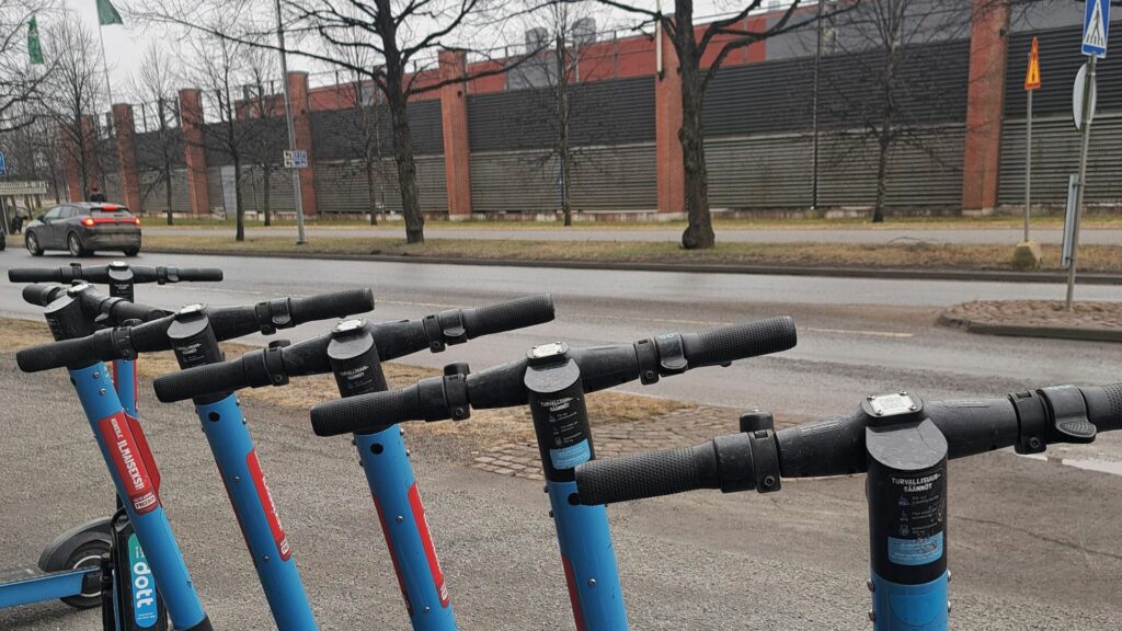 Sähköpotkulautojen ohjaustankoja rivissä, taustalla asfaltoitua ajoväylää ja pitkän rakennuksen julkisivu.