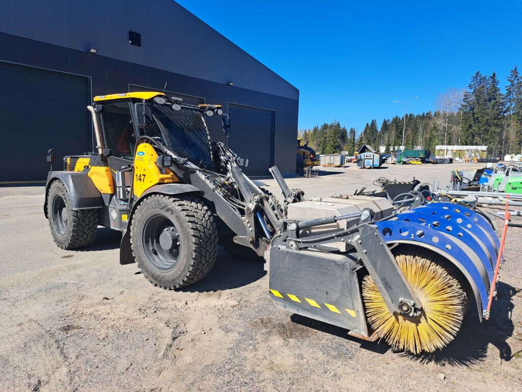 Lakaisukone pihassa, hallirakennus taustalla.