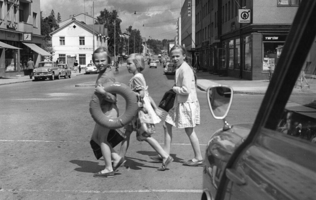 Vanha mustavalkoinen valokuva jossa kolme tyttöä ylittää suojatietoa uimarenkaat ja uimakassit käsissään.