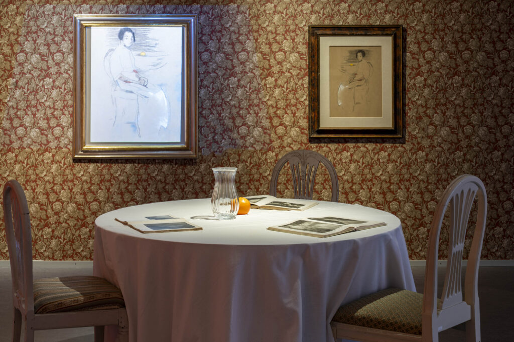 Näyttely sali jossa kukalliset tapetit ja seinillä taideteoksia. Etualalla pöytä jossa asetelma.