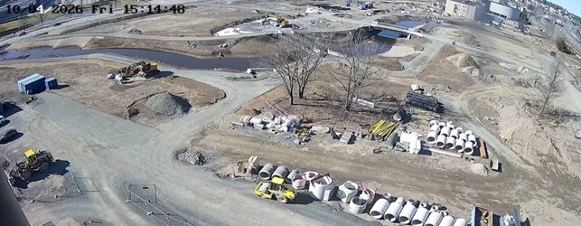 Työmaa, jossa tasaisia, juuri tehtyjä sorateitä. Pyöreitä tyhjiä betonisia rengaskaivojen pätkiä teiden varsilla. Taustalla joen uoma ja kerrostaloja.