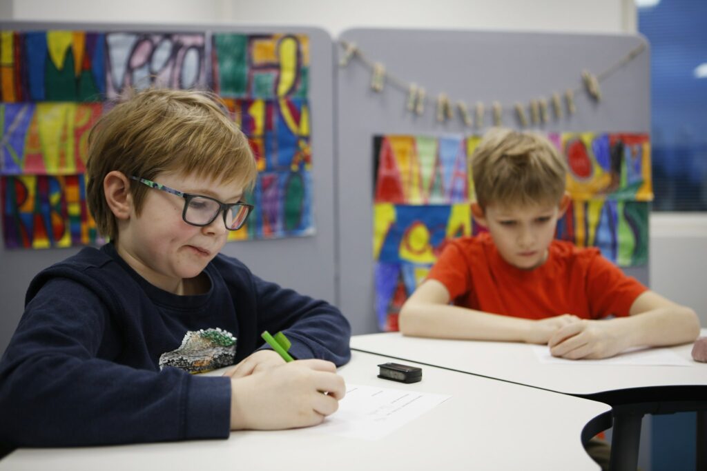 Alakouluikäinen poika istuu pulpetin ääressä suu miettiväisesti mutrulla. Hänellä on edessään pulpetilla paperi ja kynä kädessä. Taustalla luokassa toinenkin poika pulpettinsa ääressä.