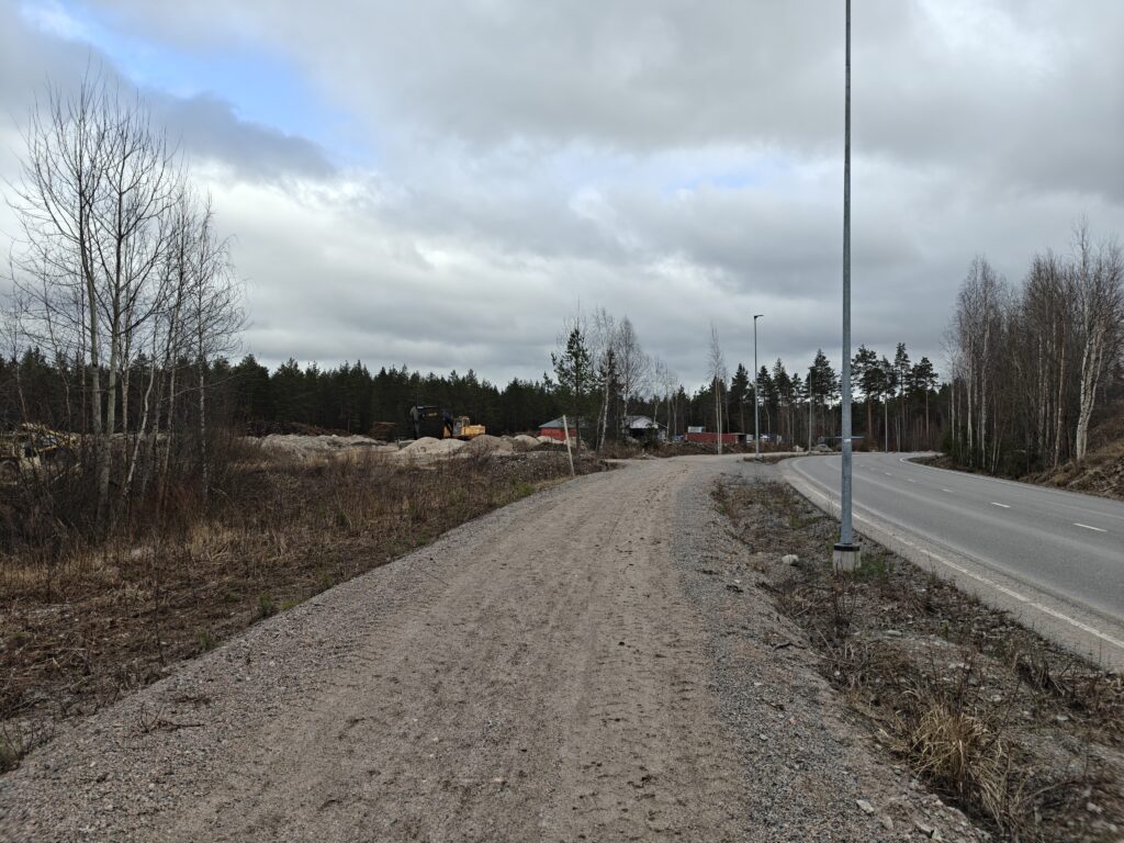 Edustalla sorapintainen jalankulkuväylä ja asfaltoitu katu, taustalla varastorakennuksia, maansiirtokone ja metsää.