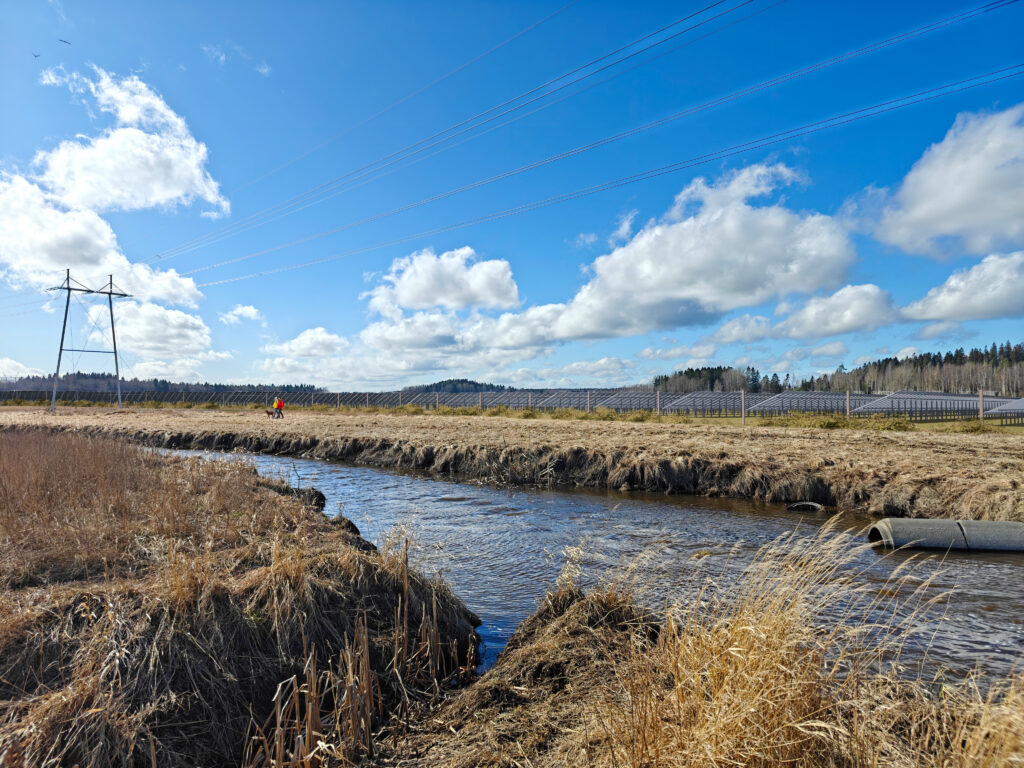 Edustalla Vantaanjoki, jonka takana avoin peltoalue. Peltoalueella näkyy aurinkopaneeleja. Kuvassa näkyy voimajohtolinja.