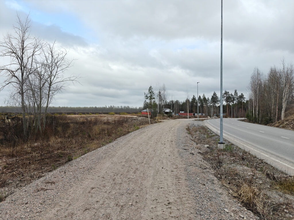 Edustalla sorapintainen jalankulkuväylä ja asfaltoitu katu, taustalla varastorakennuksia, maansiirtokone ja aurinkopaneelit. Kaukomaisemassa metsää.