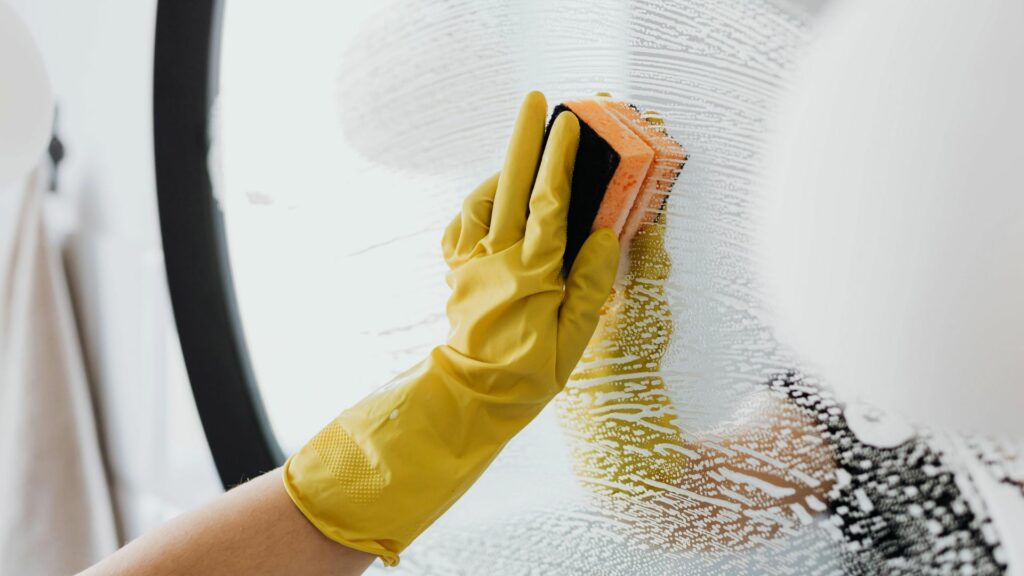 Keltaisen kumihanskan verhoama käsi pyyhkii pesusienellä pyöreää peiliä puhdistusaine vaahdoten.