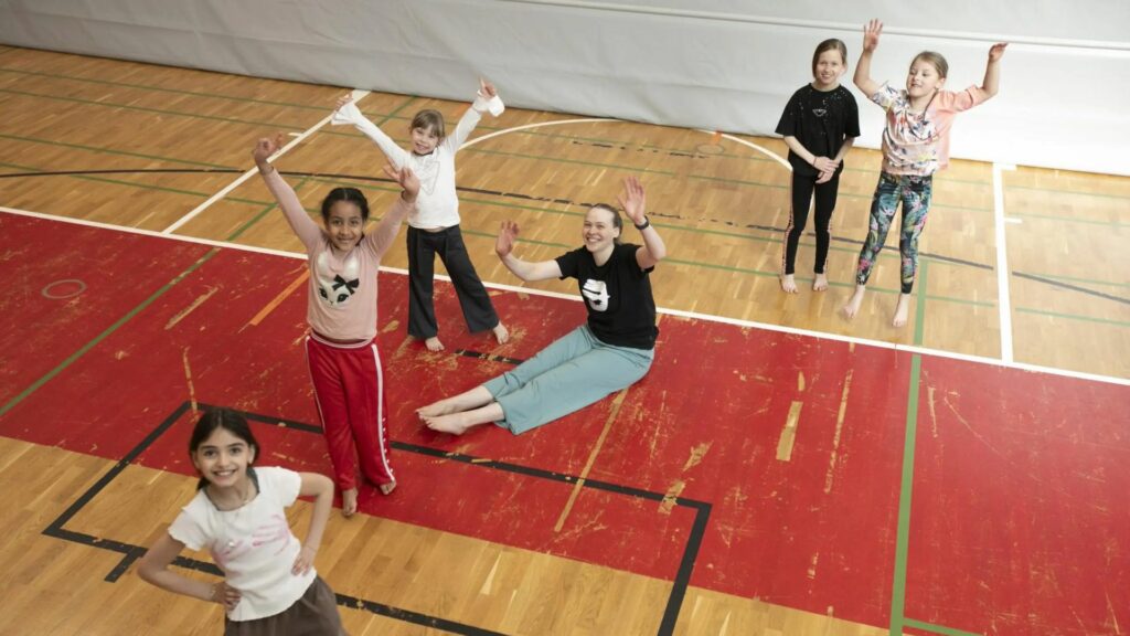 Viisi alakouluikäistä tyttöä seisomassa kädet ilmassa iloisina liikuntasalissa. Heidän joukossaan istuu opettaja, jolla myös kädet iloisesti ilmassa.