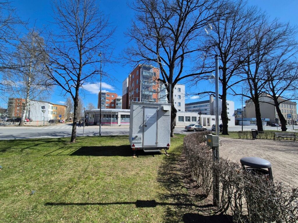 Kontti puiston nurmikolla. Taustalla kerrostaloja. Keväinen vaaleansininen taivas, puiston lehtipuissa ei ole vielä lehtiä.
