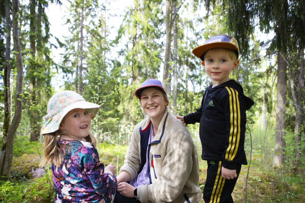 Äiti ja kaksi lasta ovat metsässä ja katsovat suoraan kameraan.