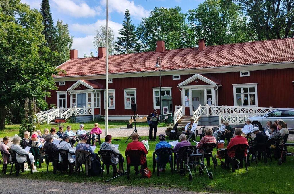 Kuvassa senioripuistoilua Antonin talon pihalla. Ihmiset istuvat ringissä ja laulaja esiintyy keskellä.