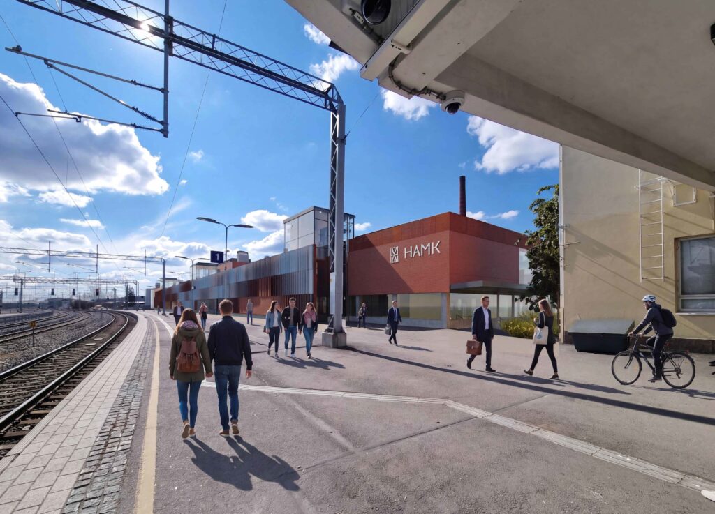 Valokuvaupote: Näkymä radan puolelta pohjoisen suunnasta kohti uutta HAMKin kampusta. Rakennus on punatiilinen. On lähes pilvetön kesäpäivä. Ihmisiä kulkee asemalaiturilla.